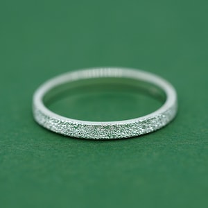 May include: A silver ring with a textured surface. The ring is set against a green background. The ring's band has a textured, glittery appearance.