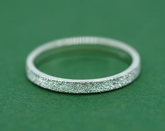 May include: A silver ring with a textured surface. The ring is set against a green background. The ring's band has a textured, glittery appearance.