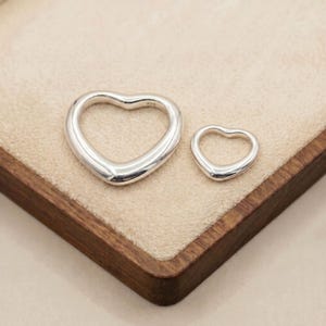 Peut inclure: Deux pendentifs en forme de cœur en argent sterling poli de tailles différentes. Le plus grand cœur porte le poinçon 925 gravé sur sa courbe supérieure droite. Les deux pièces ont une finition de surface métallique lisse et réfléchissante.