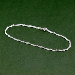 Könnte beinhalten: Ein silbernes Kettenarmband mit einem zarten, gedrehten Design. Das Armband liegt auf einem grünen Hintergrund.
