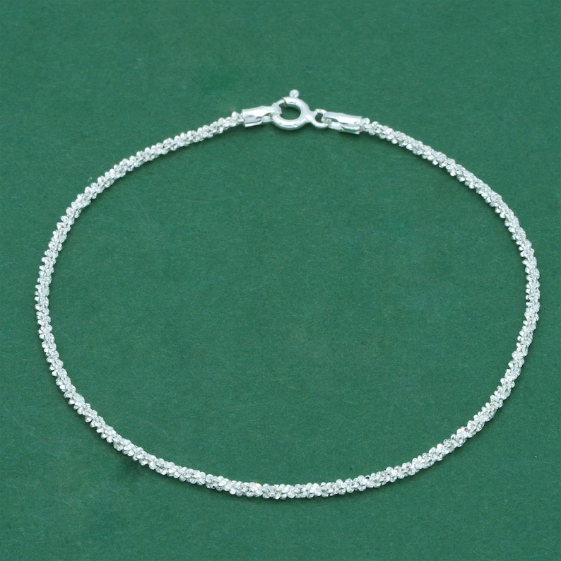 K&ouml;nnte beinhalten: Ein filigranes Silberarmband mit einem strukturierten, funkelnden Design. Das Armband verf&uuml;gt &uuml;ber einen sicheren Verschluss und wird vor einem einfarbigen gr&uuml;nen Hintergrund pr&auml;sentiert. Dieses Schmuckst&uuml;ck eignet sich f&uuml;r den Alltag oder besondere Anl&auml;sse.