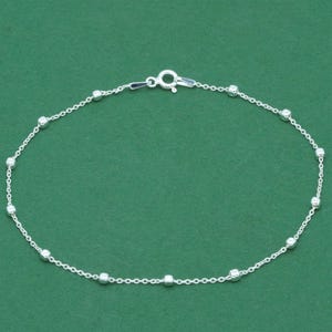 May include: A delicate silver anklet with small, round beads evenly spaced along a fine chain. The anklet features a secure clasp and is displayed against a solid green background. This piece of jewellery is suitable for everyday wear.