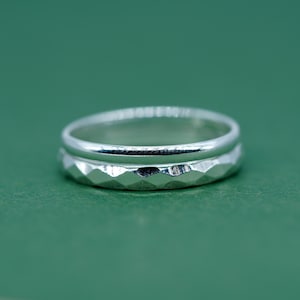 May include: Two silver rings, one on top of the other. The lower ring has a textured, faceted pattern, and the upper ring is smooth. The rings are displayed against a plain green backdrop.