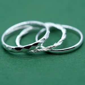 May include: Four silver rings of different designs are shown against a green backdrop. One ring has a hammered texture, another has a twisted rope design, and the third is smooth. These are likely jewellery items.