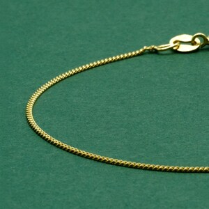 May include: Close-up of a gold-colored chain necklace with a delicate, textured design. The chain is set against a solid green background. The clasp is visible at the top right.