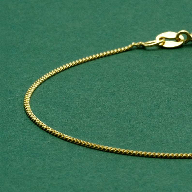 May include: Close-up of a gold-colored chain necklace with a delicate, textured design. The chain is set against a solid green background. The clasp is visible at the top right.