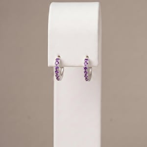 May include: A pair of small silver hoop earrings featuring a row of round, vibrant purple gemstones set along the front-facing curve of each hoop. The earrings are displayed side-by-side, showcasing the metallic finish and the purple stones.
