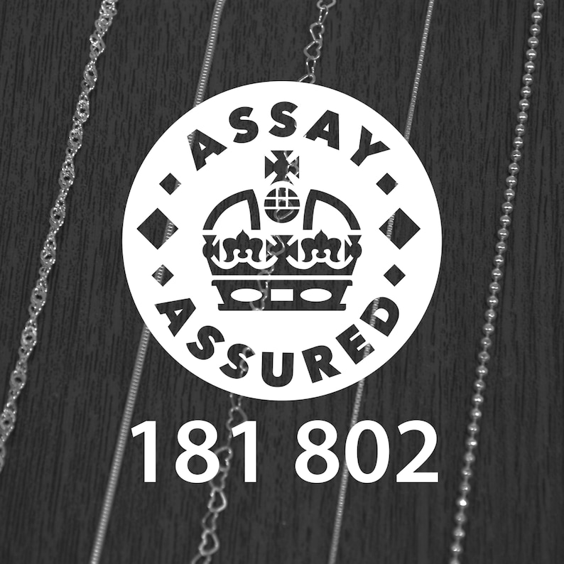 K&ouml;nnte beinhalten: Schwarz-wei&szlig;-Bild mit einem wei&szlig;en runden Emblem mit der Aufschrift "ASSAY ASSURED" und einem Kronen-Design. Darunter die Zahlen "181 802". Zarte Silberketten sind auf beiden Seiten des Emblems zu sehen.