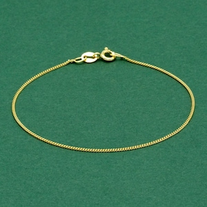 May include: A delicate gold-colored chain bracelet with a lobster clasp closure. The bracelet is displayed on a solid green background. The chain is thin and has a textured, woven appearance.