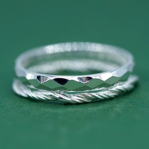 May include: Three silver rings, one smooth, one with a faceted design, and one with a twisted rope design. The rings are stacked on top of each other against a green background. The rings are made of silver.