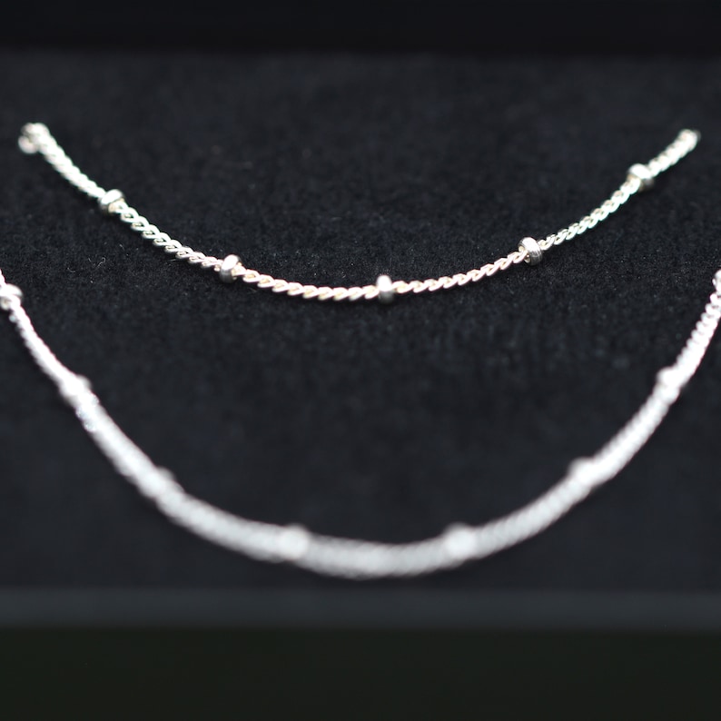 May include: Two silver necklaces with small round beads on a black background.