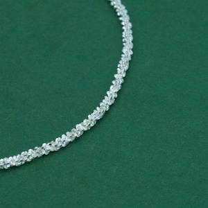 K&ouml;nnte beinhalten: Eine silberne Halskette mit einem gedrehten Seildesign. Die Halskette besteht aus kleinen, reflektierenden Perlen, die das Licht einfangen. Der Hintergrund ist einfarbig dunkelgr&uuml;n.