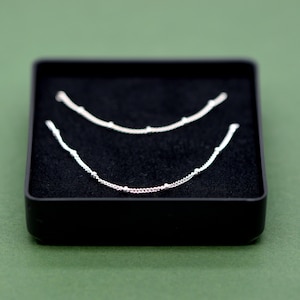 May include: Two silver chains with small beads, displayed in a black velvet box.