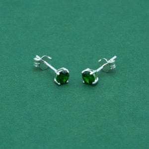 May include: A pair of silver stud earrings featuring round, vivid green gemstones. The earrings have a simple, classic design, suitable for daily wear. The gemstones are a deep, emerald green colour.