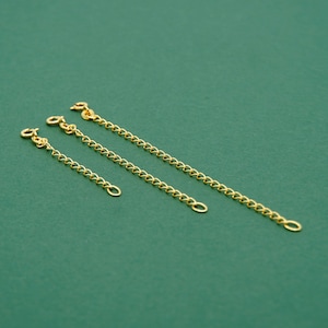 May include: Three gold-coloured chain extenders of varying lengths are arranged diagonally on a green background. Each extender has a lobster clasp on one end and a small loop on the other, designed to lengthen necklaces or bracelets.