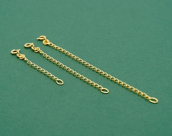 May include: Three gold-colored chain extenders of varying lengths are arranged diagonally on a green background. Each extender has a lobster clasp on one end and a small loop on the other, designed to lengthen necklaces or bracelets.