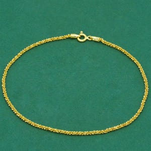 May include: A fine gold bracelet with a textured, beaded appearance. The bracelet is set against a plain green backdrop. The clasp is visible, and the bracelet seems to be made of small, connected beads.