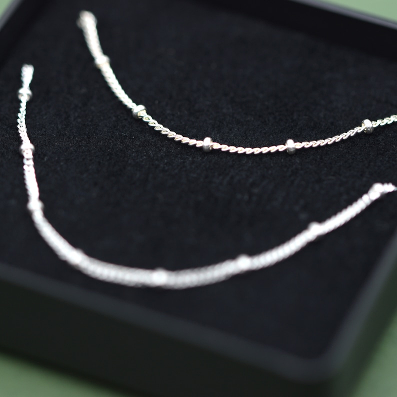 May include: Two silver necklaces with small beads on a black velvet background.
