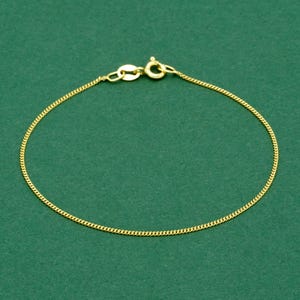 May include: A delicate gold-colored chain bracelet. The bracelet features a fine curb chain design with a lobster clasp closure. The jewelry is displayed against a solid green background.