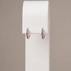 May include: A pair of small silver hoop earrings featuring a row of embedded round-cut pink gemstones. The metal has a polished finish, and the stones are set along the front-facing curve of each hoop, creating a consistent and symmetrical appearance.