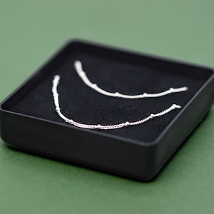 May include: Two silver necklaces with small beads on a black velvet background inside a black box.