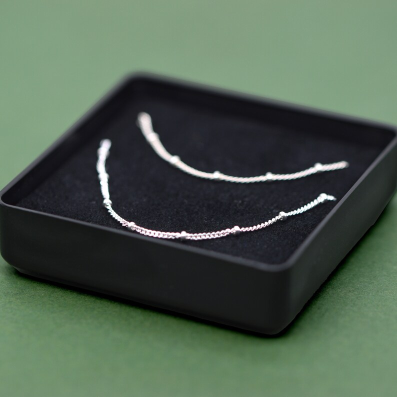 May include: Two silver necklaces with small beads on a black velvet background inside a black box.