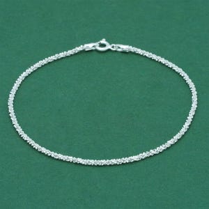 K&ouml;nnte beinhalten: Ein filigranes Silberarmband mit einem strukturierten, perlenbesetzten Design. Das Armband wird vor einem einfarbigen gr&uuml;nen Hintergrund pr&auml;sentiert, der seine komplizierten Details und den sicheren Verschluss hervorhebt.