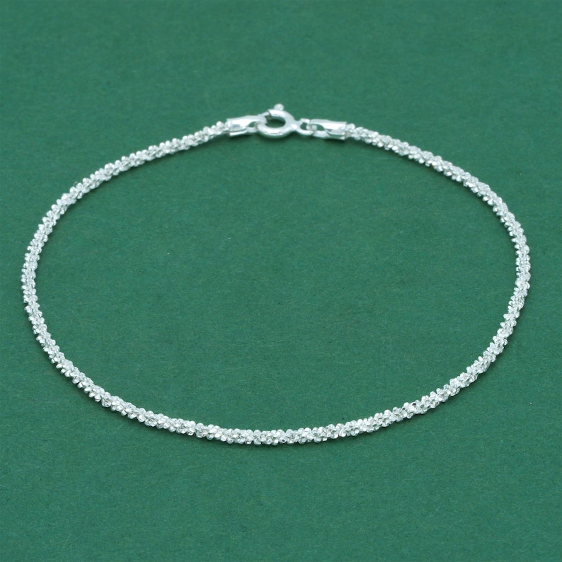 K&ouml;nnte beinhalten: Ein filigranes Silberarmband mit einem strukturierten, perlenbesetzten Design. Das Armband wird vor einem einfarbigen gr&uuml;nen Hintergrund pr&auml;sentiert, der seine komplizierten Details und den sicheren Verschluss hervorhebt.