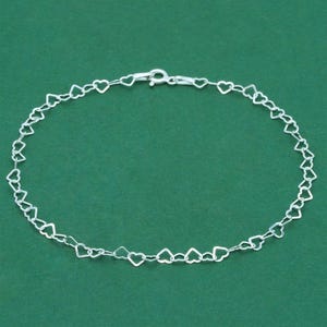 May include: A delicate silver bracelet featuring a repeating heart design. The bracelet is composed of small, interconnected heart-shaped links, creating a charming and romantic accessory. The clasp is visible at the top, and the bracelet is displayed against a green background.