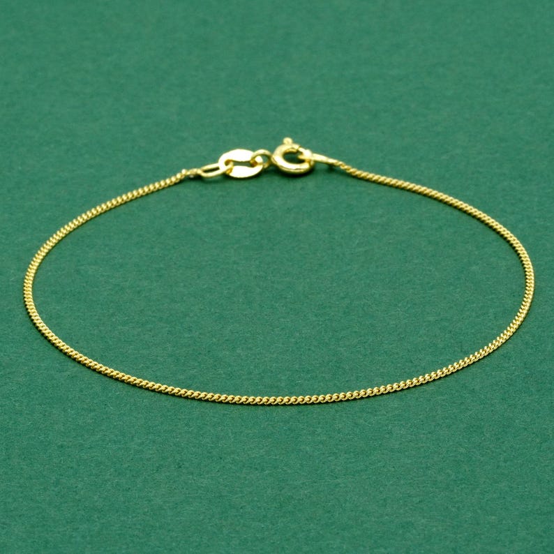 May include: A delicate gold-colored bracelet with a curb chain design. The bracelet features a lobster clasp closure and is displayed against a solid green background. The chain is approximately 7 inches in length.