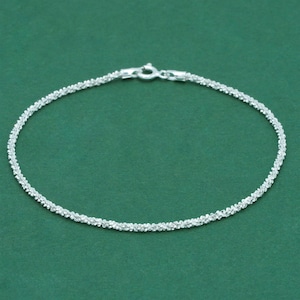 K&ouml;nnte beinhalten: Ein filigranes Silberarmband mit einem strukturierten, perlenbesetzten Design. Das Armband hat einen kleinen Verschluss f&uuml;r sicheres Tragen. Der Schmuck wird vor einem einfarbigen gr&uuml;nen Hintergrund pr&auml;sentiert.