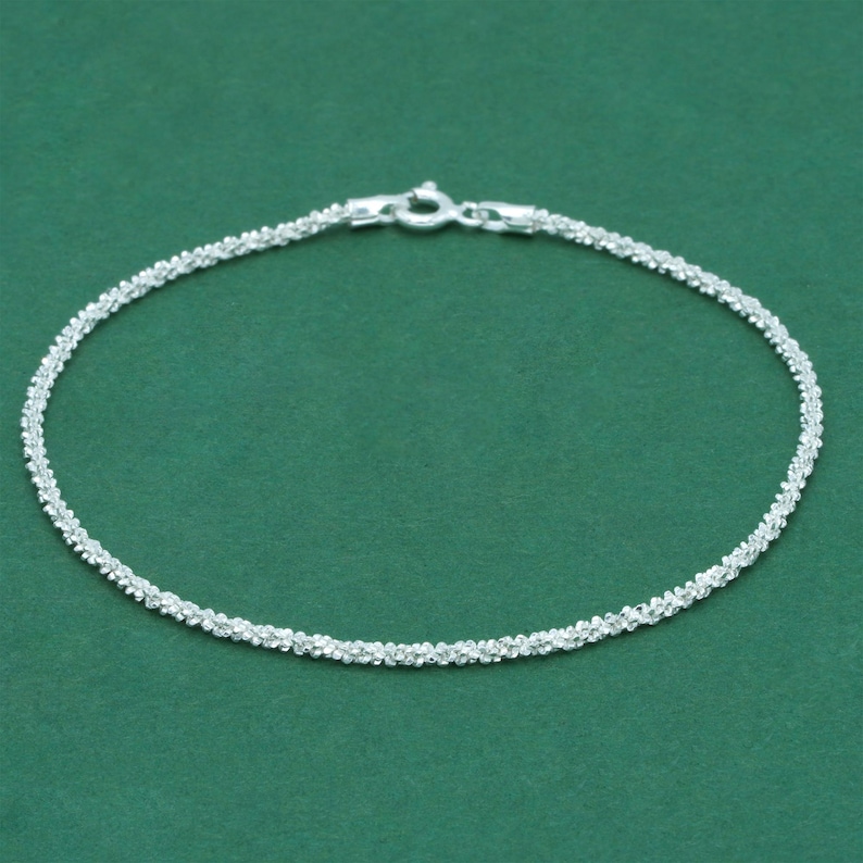 K&ouml;nnte beinhalten: Ein filigranes Silberarmband mit einem strukturierten, perlenbesetzten Design. Das Armband hat einen kleinen Verschluss f&uuml;r sicheres Tragen. Der Schmuck wird vor einem einfarbigen gr&uuml;nen Hintergrund pr&auml;sentiert.