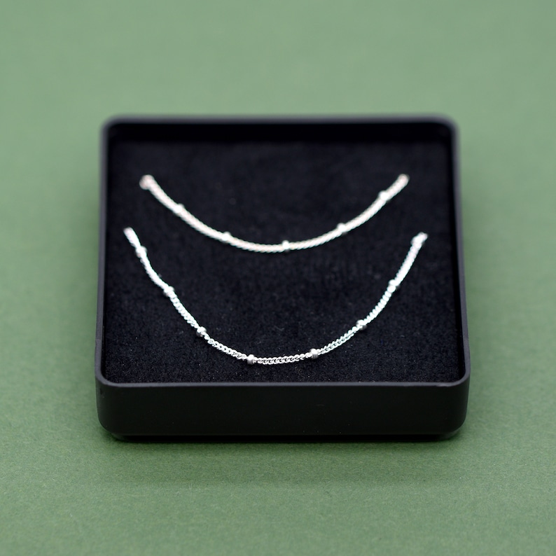 May include: Two silver necklaces with small beads on a black velvet background.