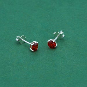 May include: A pair of silver stud earrings, each featuring a bright red gemstone. The earrings are set against a green backdrop, highlighting their colour and design. The gemstones are round and faceted.