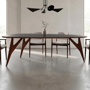 Op de afbeelding: Een moderne eethoekset met een donkere ovale tafel met houten poten en zes bijpassende stoelen. De stoelen hebben lichtgekleurde kussens. Een vaas met een plant en een zwarte kroonluchter maken de scène compleet.
