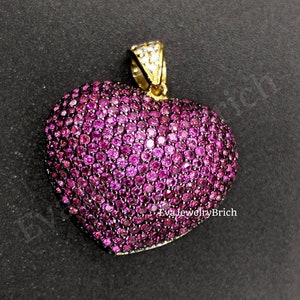 May include: A heart-shaped pendant, covered in small, bright pink gemstones. The pendant has a gold-coloured bail at the top for a chain. The gemstones are set closely together, creating a sparkling effect. The background is a dark, solid colour.