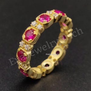 Può includere: Un anello color oro con un motivo ripetuto di pietre preziose rosa brillante e piccole pietre bianche. L'anello ha una fascia strutturata e uno sfondo scuro.
