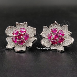 Op de afbeelding: Een paar bloemvormige oorknopjes met een cluster van ovaal geslepen levendige roze stenen in het midden, omringd door bloemblaadjes bezet met talloze kleine heldere witte steentjes. De tekst EvaJewelryBrich is in het midden zichtbaar.