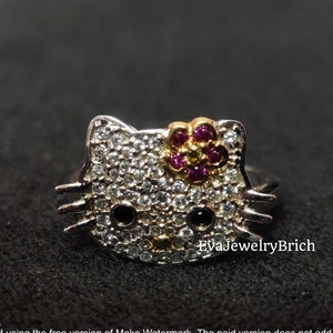 May include: A silver ring with a white, black, and pink Hello Kitty design. The ring is covered in small, clear rhinestones. The Hello Kitty design has a pink flower on top with a yellow center.