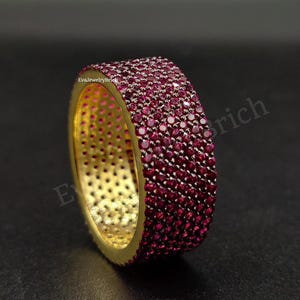 Peut inclure: Une bague dorée entièrement recouverte de petites pierres précieuses rondes et rouge foncé. Les pierres précieuses créent une surface continue et scintillante sur le corps de la bague. L'intérieur de la bague est de couleur or contrastante.