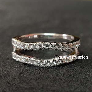 Könnte beinhalten: Ein silberner Ring mit einem geschwungenen Design, der auf jeder Seite eine Reihe kleiner Diamanten aufweist. Der Ring ist so konzipiert, dass er als Trauringverstärker getragen werden kann.