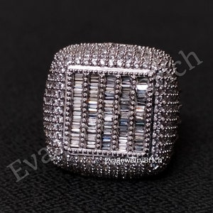 Puede incluir: Un anillo de forma cuadrada con una piedra grande rectangular en el centro rodeada de piedras más pequeñas. El anillo es de plata y tiene un acabado pulido. La piedra central es un diamante de talla baguette, y las piedras más pequeñas son diamantes de talla brillante redonda.