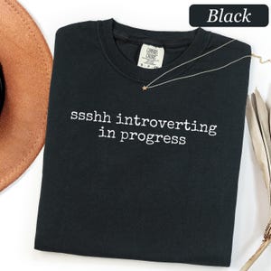 May include: A black crew neck t-shirt featuring the white text "ssshh introverting in progress" printed across the chest. The shirt has a visible brand label inside the collar and is displayed with a thin gold-toned necklace with a small star charm.