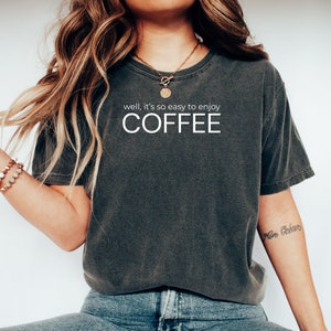 May include: A dark gray t-shirt with the text "well, it's so easy to enjoy COFFEE" printed on the front.