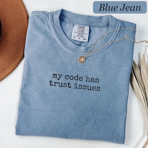以下が含まれることがあります： クルーネックと半袖のブルーデニムカラーのTシャツ。胸の中央には「my code has trust issues」という黒い刺繍のテキストが施されています。小さな円形のペンダントが付いたゴールドトーンのネックレスが襟元に置かれています。