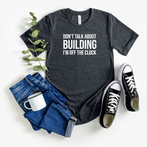 May include: A dark heather grey t-shirt with the text "Don't talk about building I'm off the clock" in white. The shirt is laid flat on a white surface with a pair of black trainers, a mug, and a pair of blue jeans.