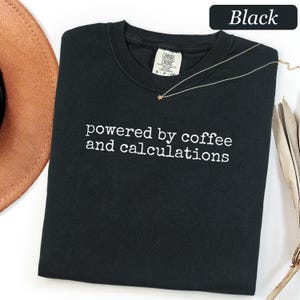 May include: A black crewneck t-shirt featuring the white embroidered text "powered by coffee and calculations" centred on the chest. The shirt has a Comfort Colors brand label at the inner neckline and a thin silver chain necklace resting on top.