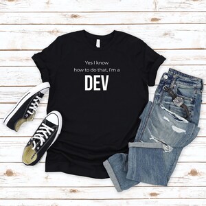 May include: Black t-shirt with white text that reads "Yes I know how to do that, I'm a DEV". The shirt is laid flat on a wood surface with a pair of black sneakers and blue jeans.