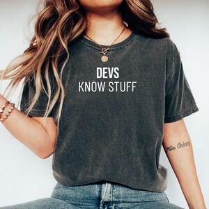 May include: A dark grey t-shirt with the text "DEVS KNOW STUFF" printed in white on the front. The shirt is being worn by a person with long brown hair and a gold necklace.
