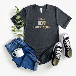 May include: A dark heather grey t-shirt with the text "Me, a DEV? I know, right?" in colorful letters. The shirt is laid out on a white surface with a pair of black sneakers, a mug, and a pair of blue jeans.